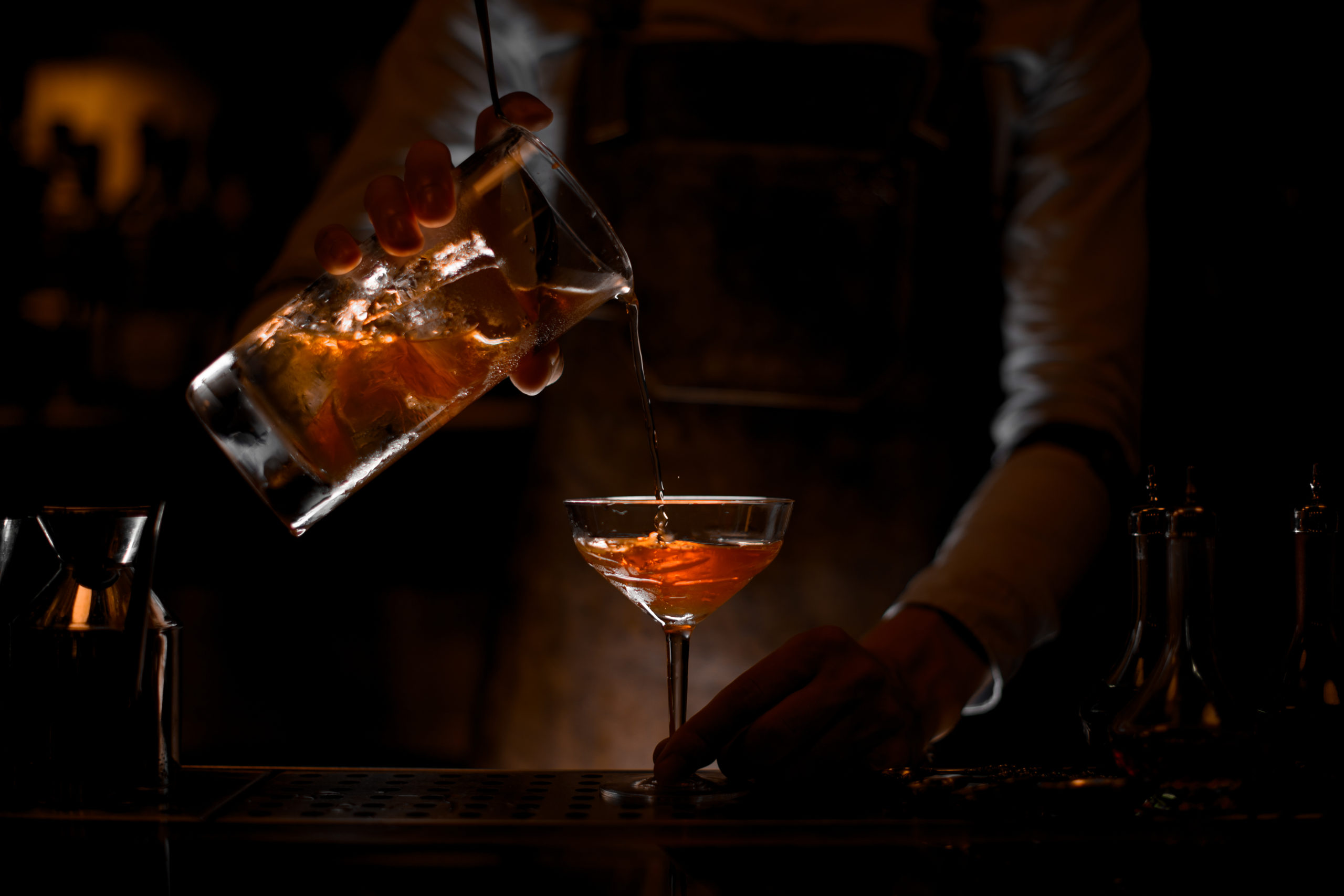Male bartender pouring a brown alcoholic cocktail from the measuring cup to the glass in the dark